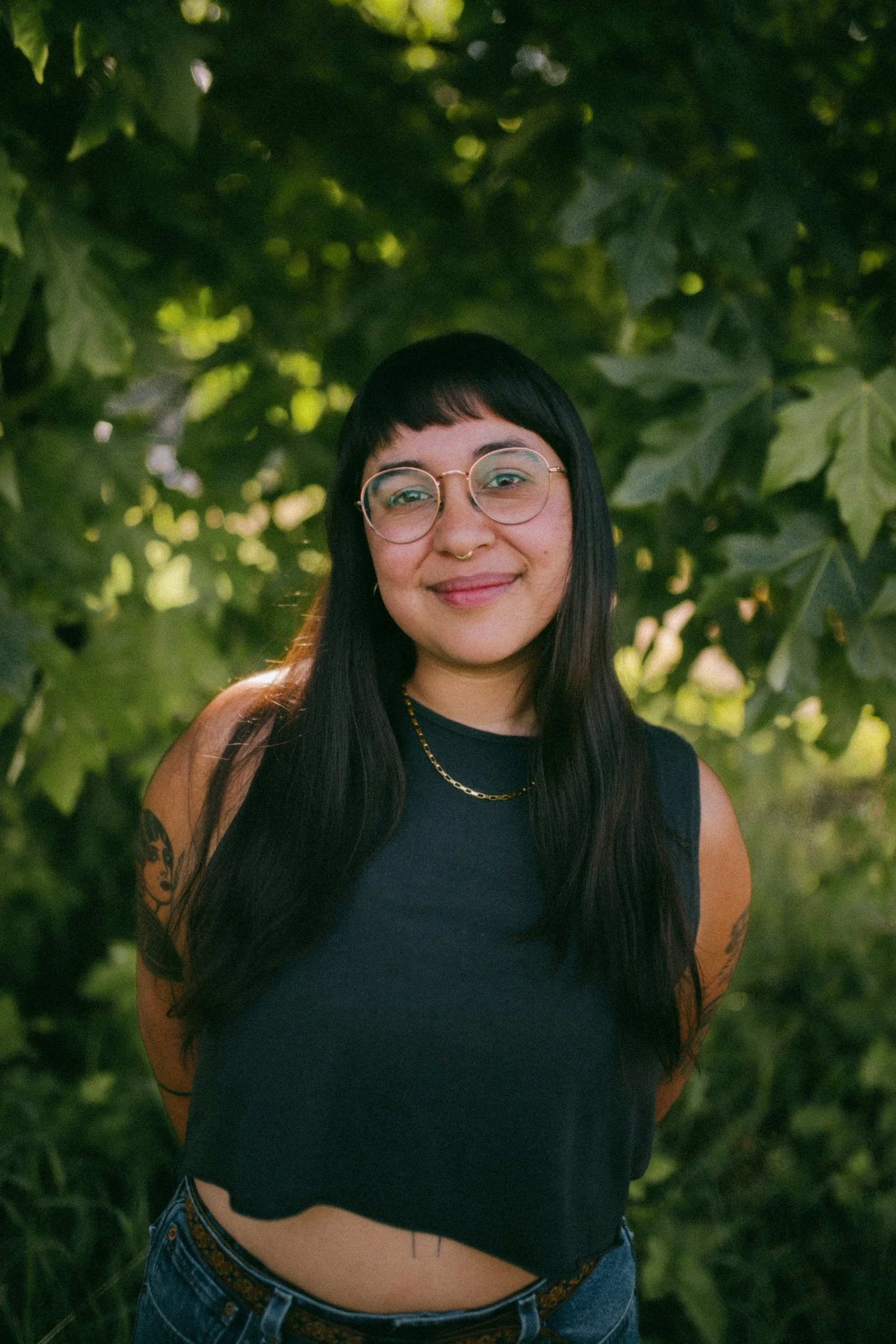 A young person with long black hair, glasses, and tattoos on her arms, standing outdoors in front of green leafy trees, smiling at the camera.