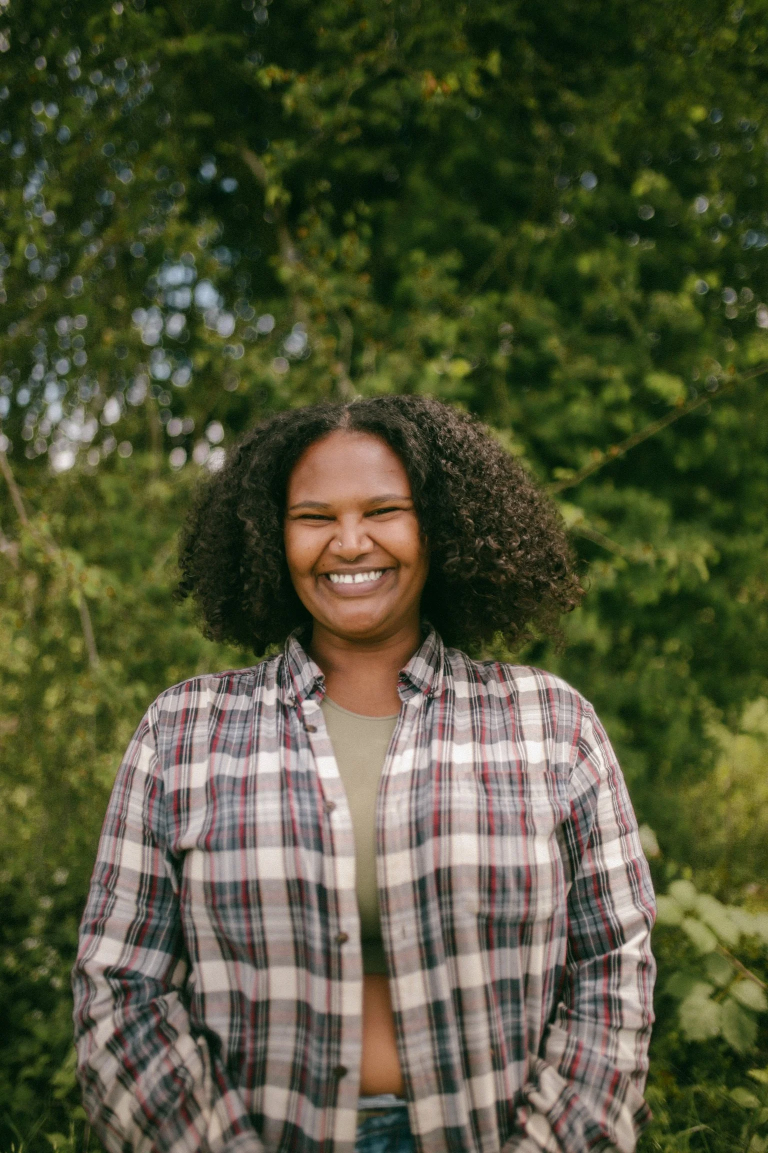 Smiling person with curly hair standing outdoors with green trees and foliage in the background.