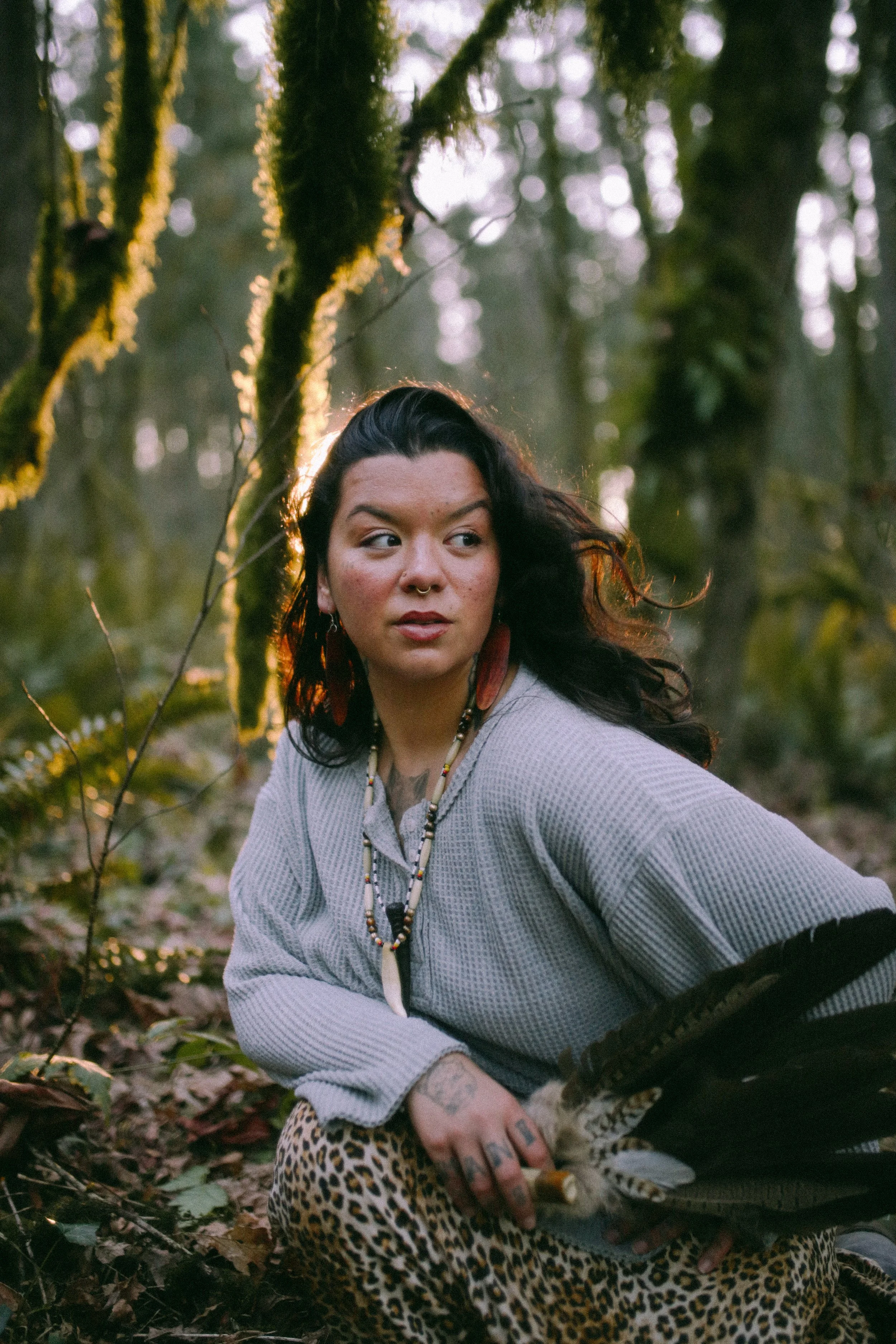 A woman with long dark hair, wearing a gray sweater, leopard print pants, and large earrings, sitting in a forest with moss-covered trees, holding a feathered fan, illuminated by the setting sun.