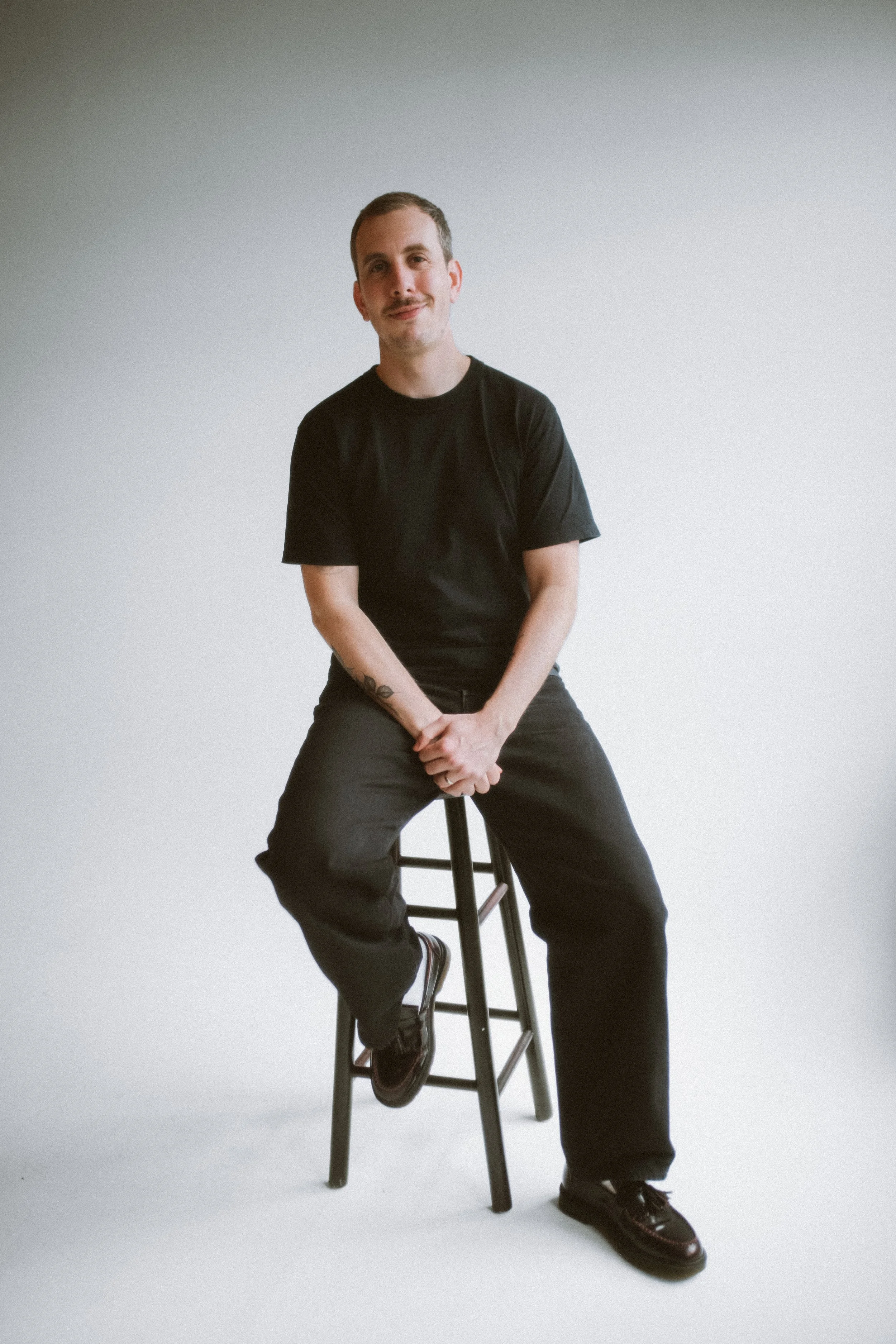 A person sitting on a black stool against a plain white background, wearing a black t-shirt, black pants, and black shoes, looking at the camera with a slight smile.