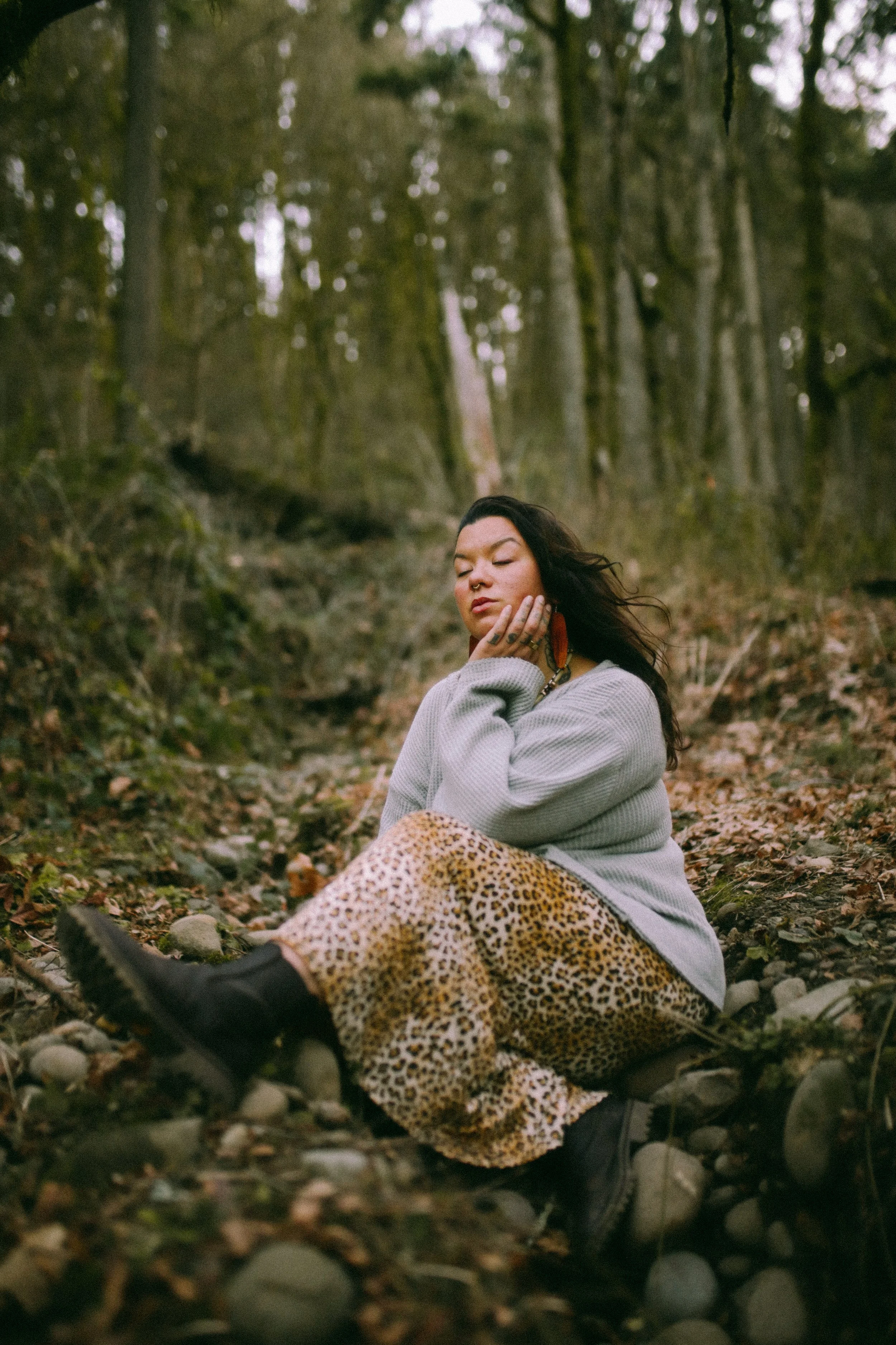 A person sitting cross-legged on a rocky forest floor with their eyes closed and hand resting on their face, surrounded by trees and fallen leaves.