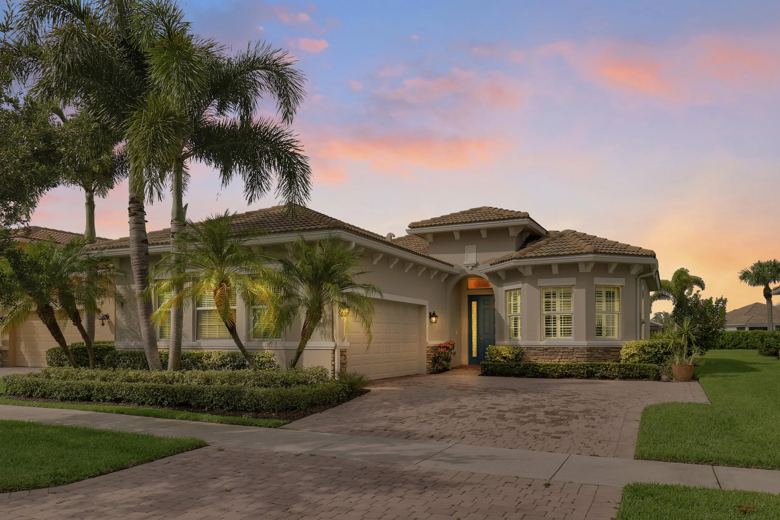 A modern two-story house with a stucco exterior, tiled roof, and large windows, surrounded by palm trees and a well-maintained lawn during sunset.