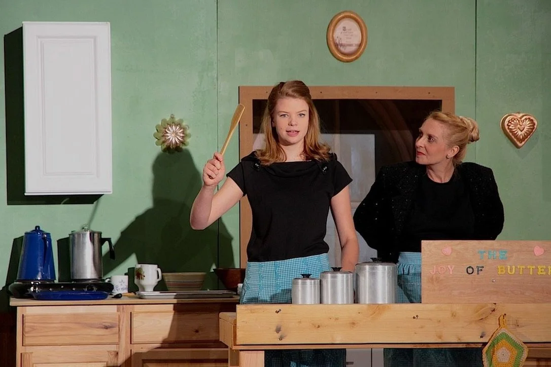 A young woman with red hair and a black shirt holding a wooden spatula, standing behind a kitchen counter, with an older woman with blonde hair and a black sweater watching her, in a kitchen with green walls and various kitchen items.