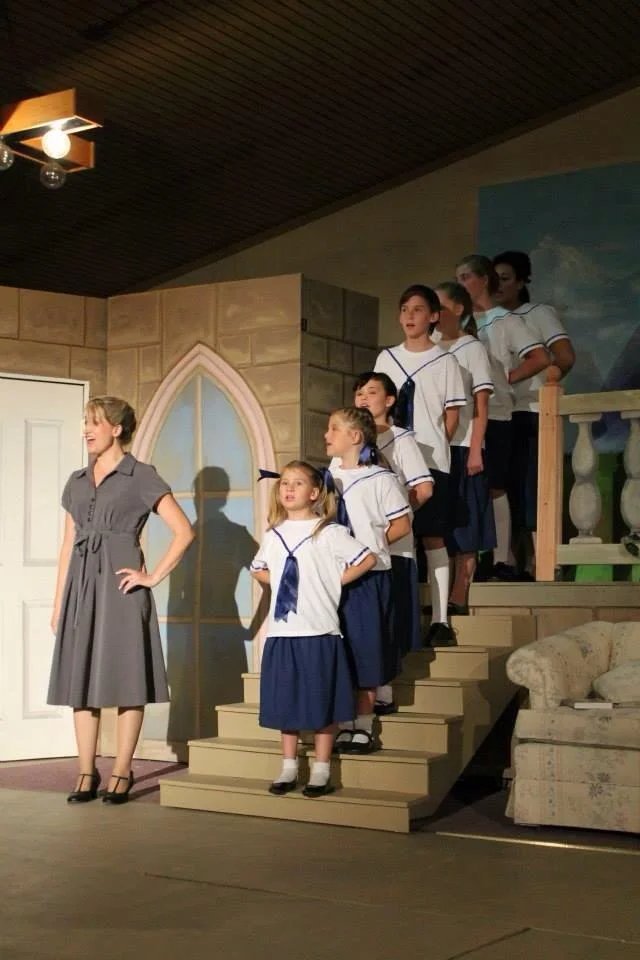 Scene from a stage play with a woman in a gray dress standing on the stage, and eight children in school uniforms standing on stairs behind her, in a set resembling a house interior.
