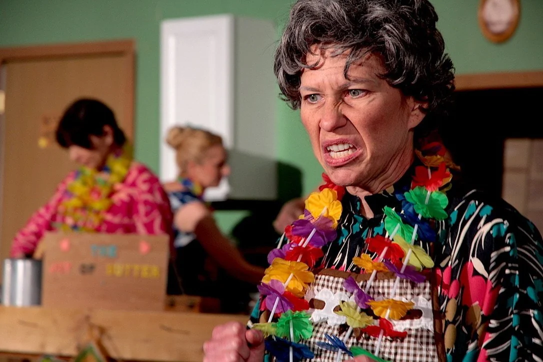 A woman with short curly hair wearing a black and white patterned shirt with pink and red accents, making an angry face. She has colorful leis around her neck and a bunch of small umbrellas. In the background, two women are decorating a cake, one wea