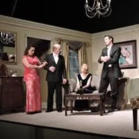 Four people in a formal living room, with three standing and one sitting. The woman on the left is wearing a red dress, the man next to her is in a dark suit, the woman seated at the table has blonde hair and is wearing a black outfit, and the man on