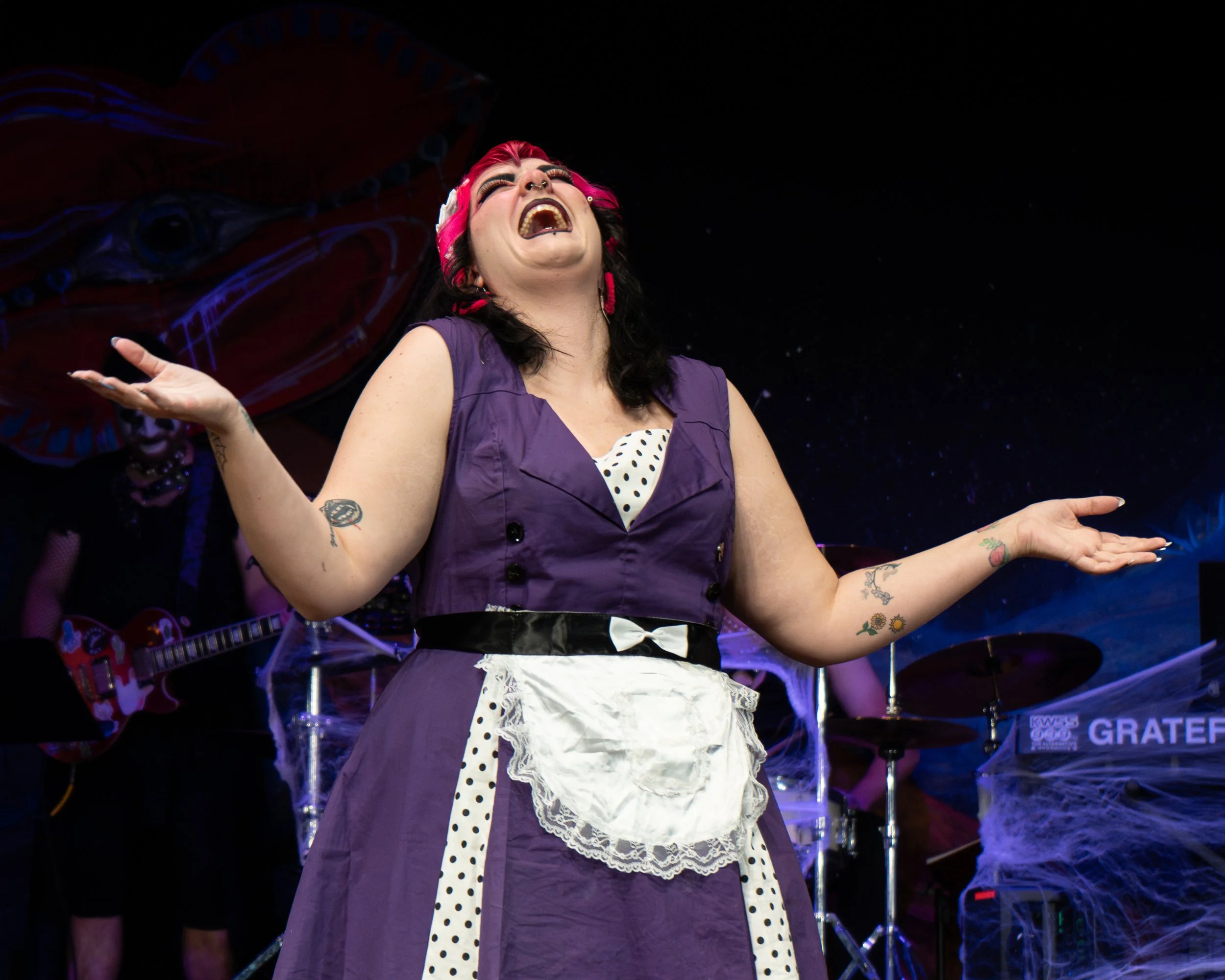 A woman with dark hair and a pink headband, wearing a purple dress with a white apron, is singing passionately on stage. She has tattoos on her arms and is gesturing with her hands. A musician playing a guitar is visible in the background, along with