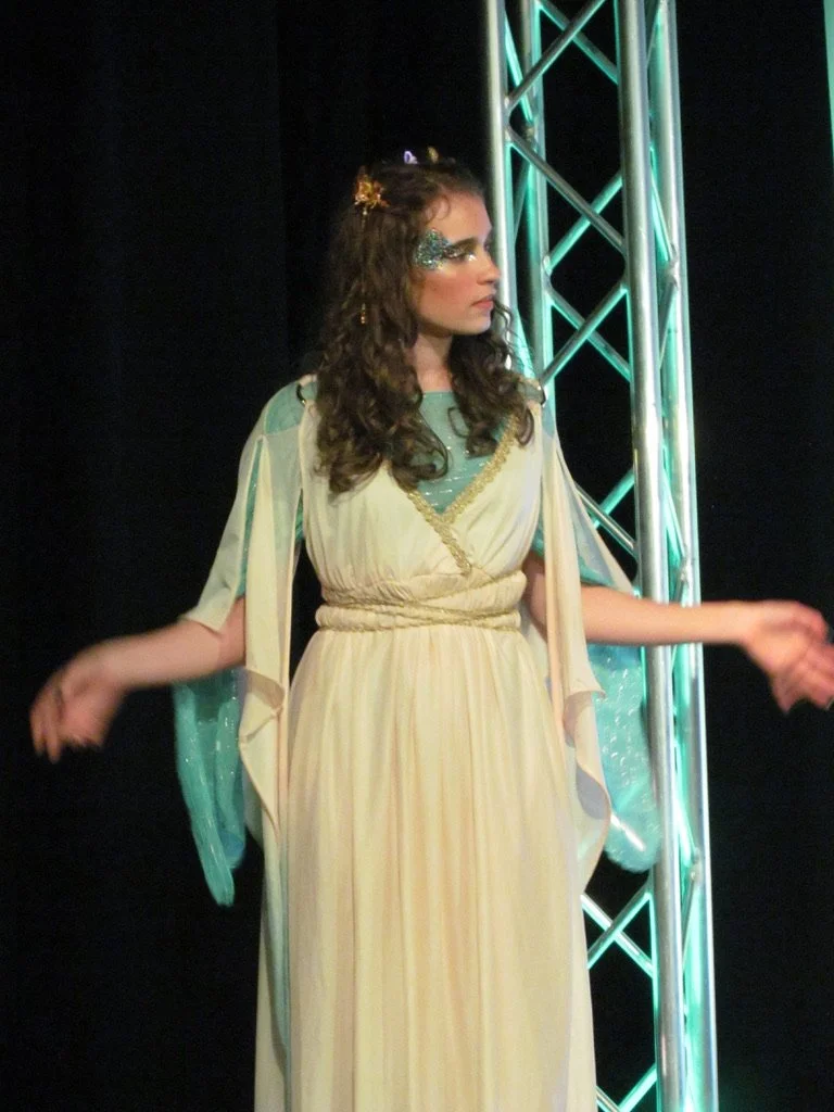 Young woman dressed as a fairy or goddess in a flowing cream and teal gown with glittery makeup, standing on stage with a metallic truss structure illuminated in teal lights.