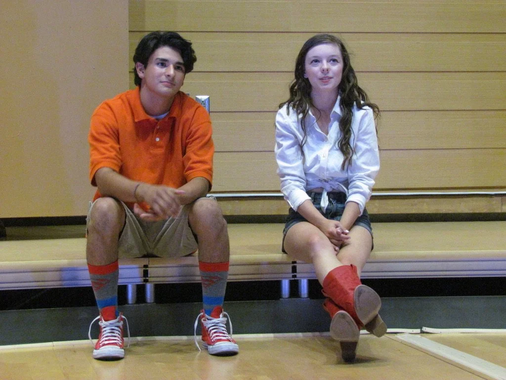 A boy and girl sitting on a wooden stage, facing forward. The boy is wearing an orange polo shirt, beige shorts, colorful striped socks, and sneakers. The girl is wearing a white blouse, dark shorts, and red boots.