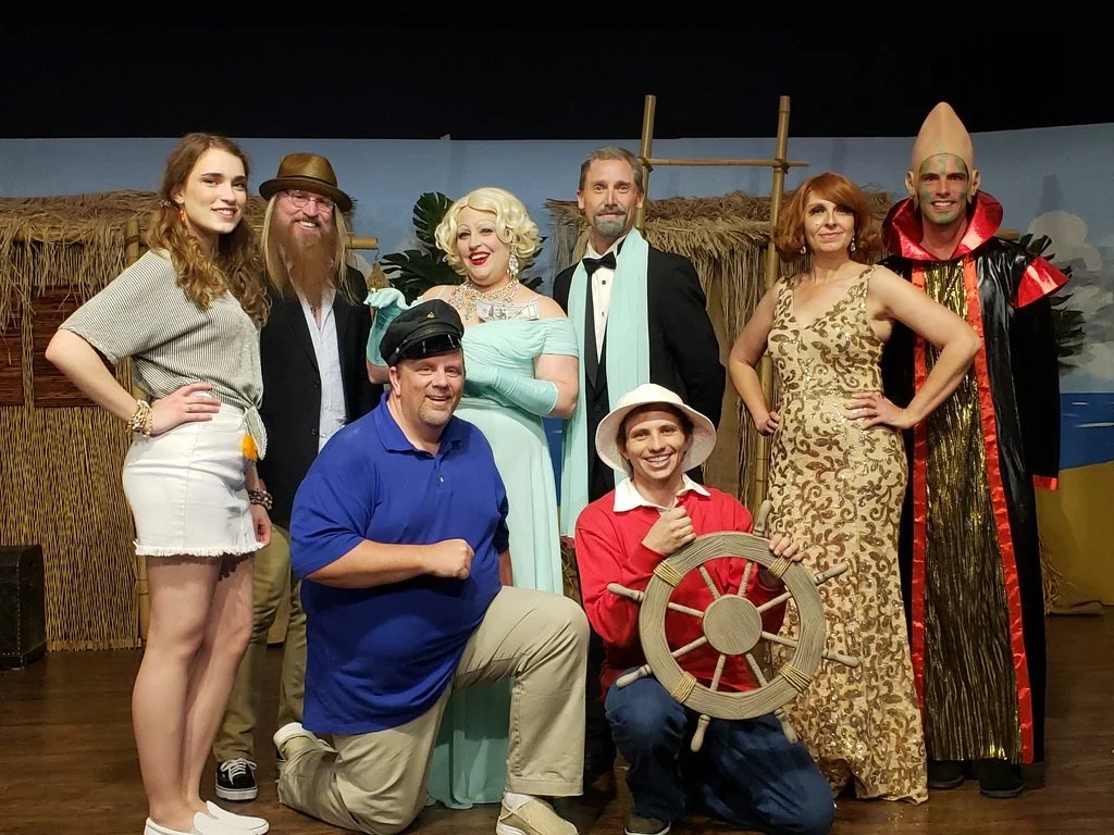 Group of seven actors in colorful costumes on a stage with a tropical backdrop, portraying a scene from a play or musical.