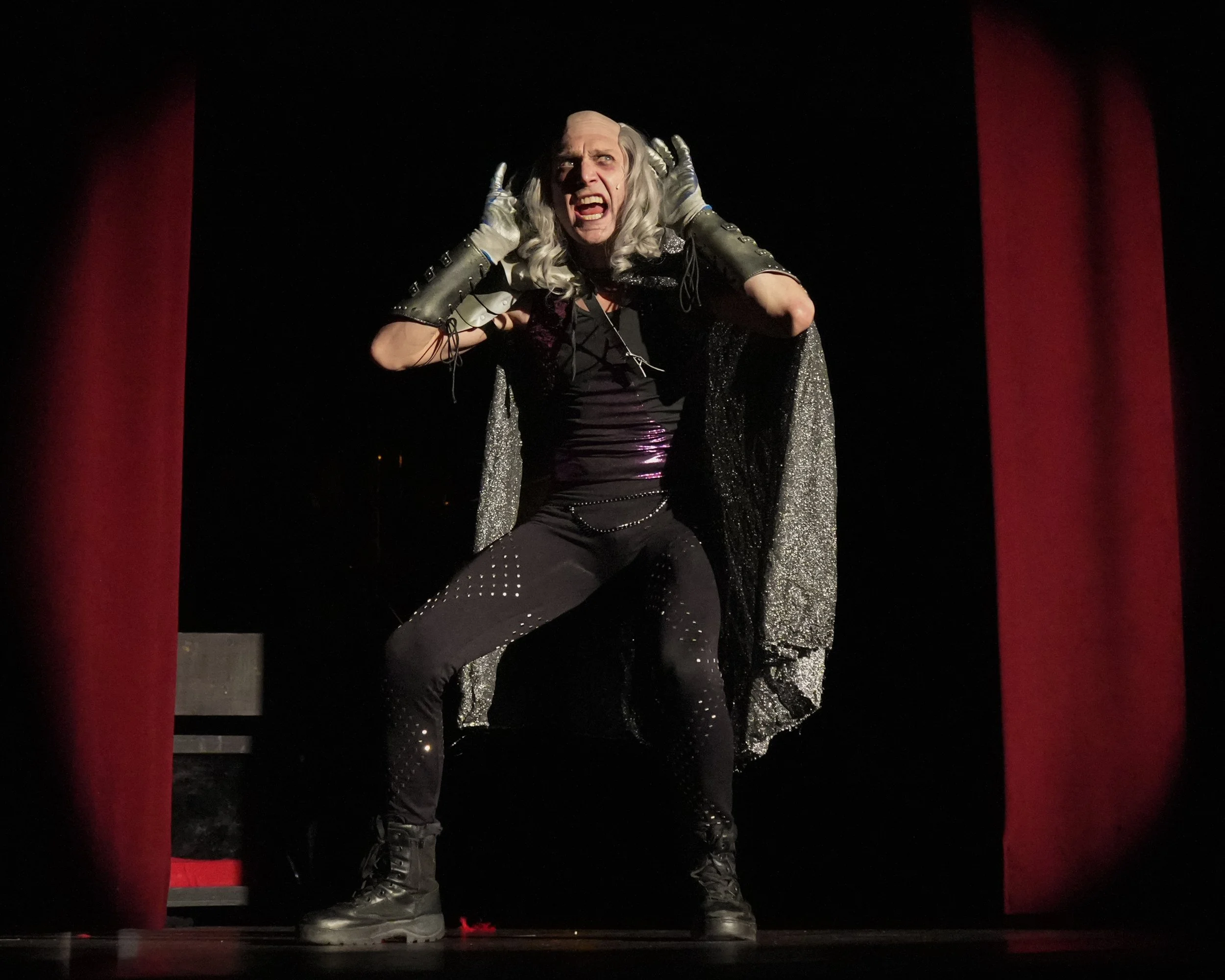 Performer dressed in black with silver hair, black boots, and a glittery cape, standing on stage between red curtains with an intense expression.