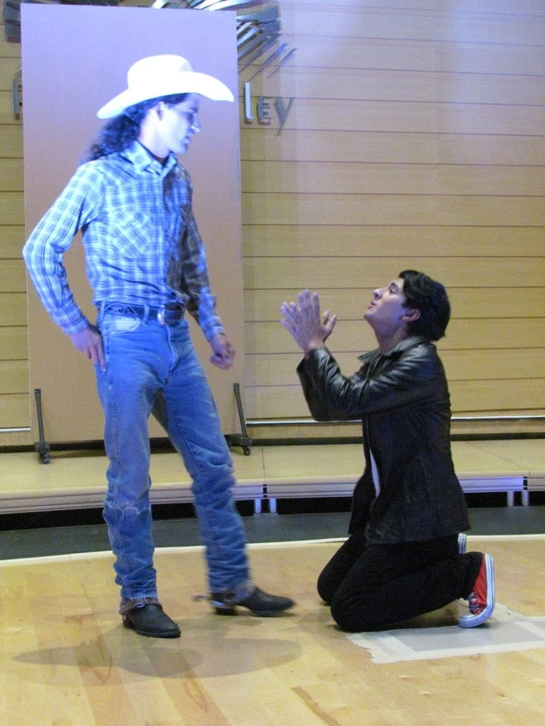 A person wearing a cowboy hat, plaid shirt, and jeans stands with hands on hips while another person kneels on the floor looking up with hands clasped in front, in a room with wood paneling.