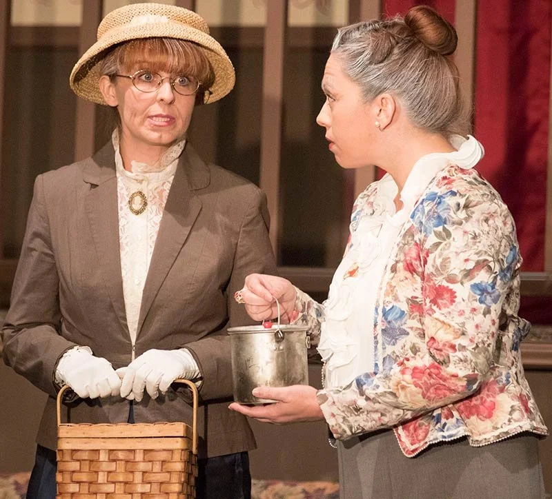 Two women dressed in vintage clothing having a serious conversation, one holding a small metal bucket and the other a wicker basket, in an indoor setting.