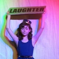A young girl standing in front of a pink curtain, holding a wooden sign that says 'LAUGHTER' above her head.