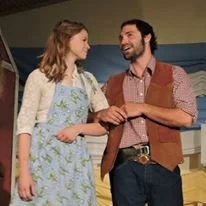 A man and woman standing and talking indoors, woman wearing a floral dress and woman wearing a plaid shirt and brown vest.