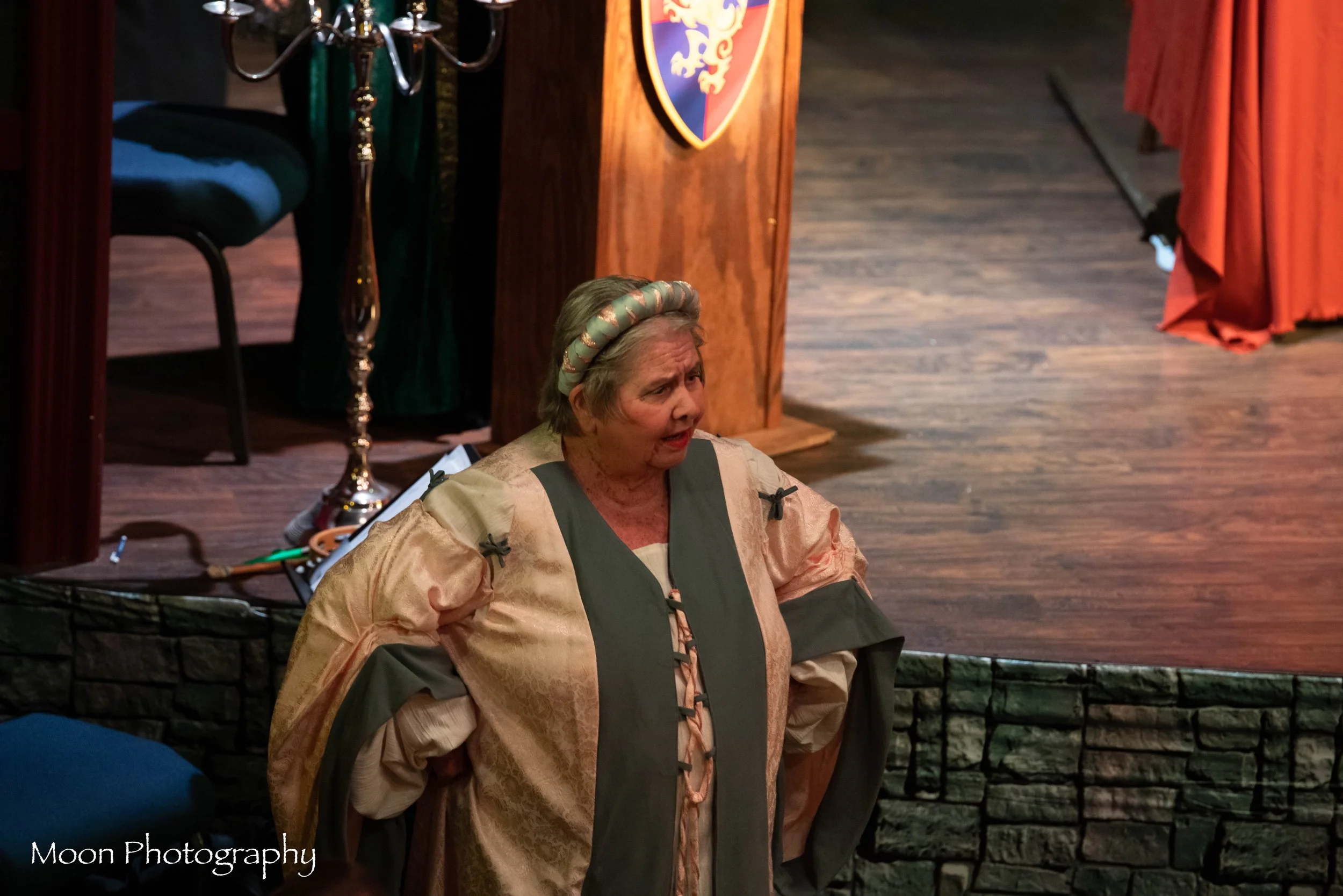 An elderly woman dressed in medieval-style clothing with wide sleeves and a headband, standing on a stage with her hands on her hips, in front of a wooden podium with a shield emblem and a cloth-covered table.