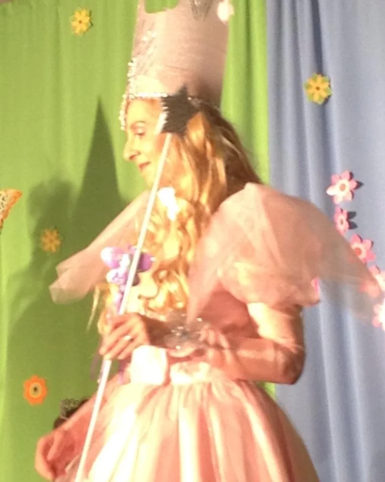 A young girl dressed as a fairy or princess, holding a magic wand, with a colorful backdrop decorated with paper flowers and a green curtain.
