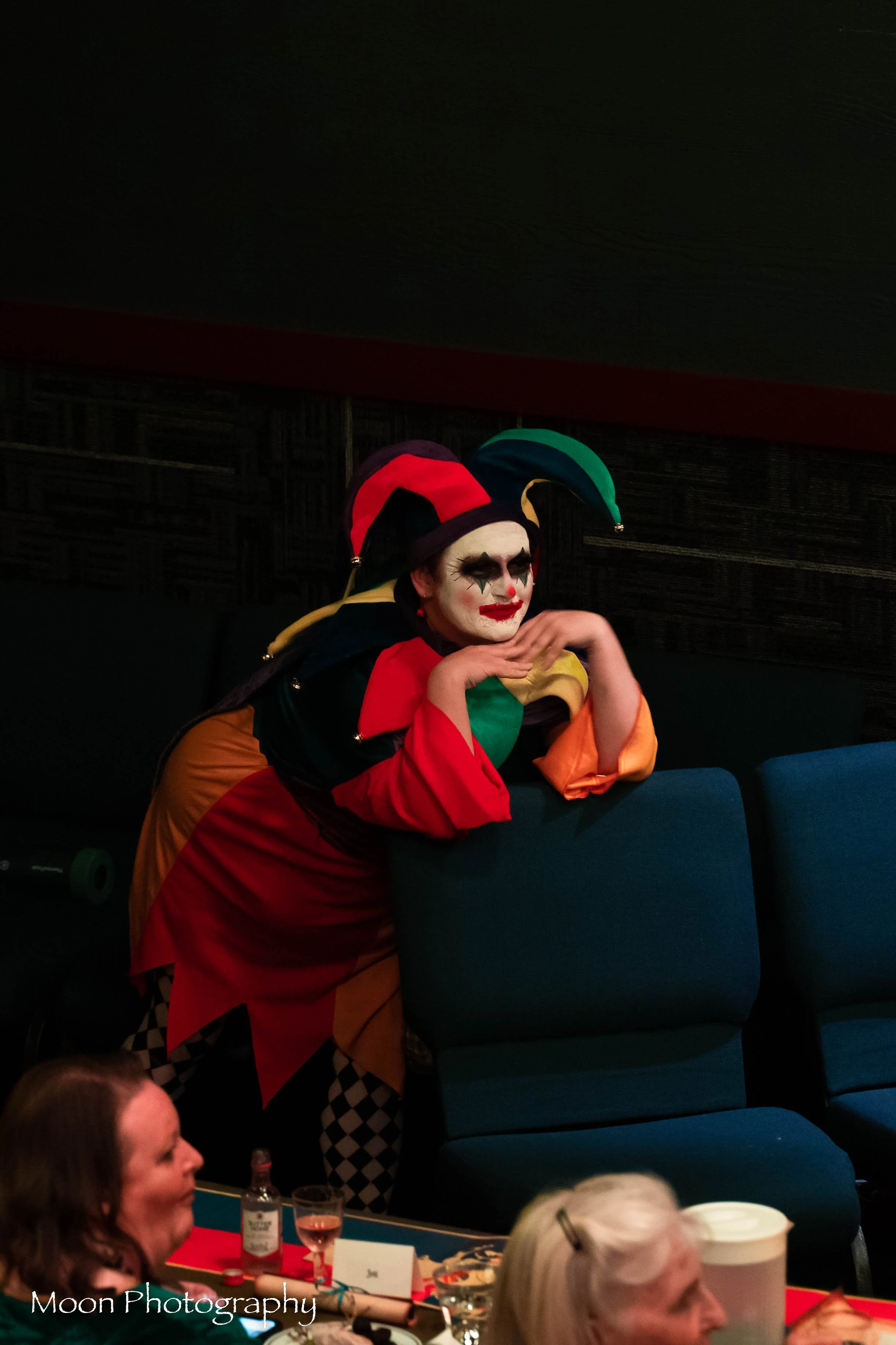 Person dressed as a jester clown with colorful costume and makeup, leaning on chairs at a table, at an indoor event.