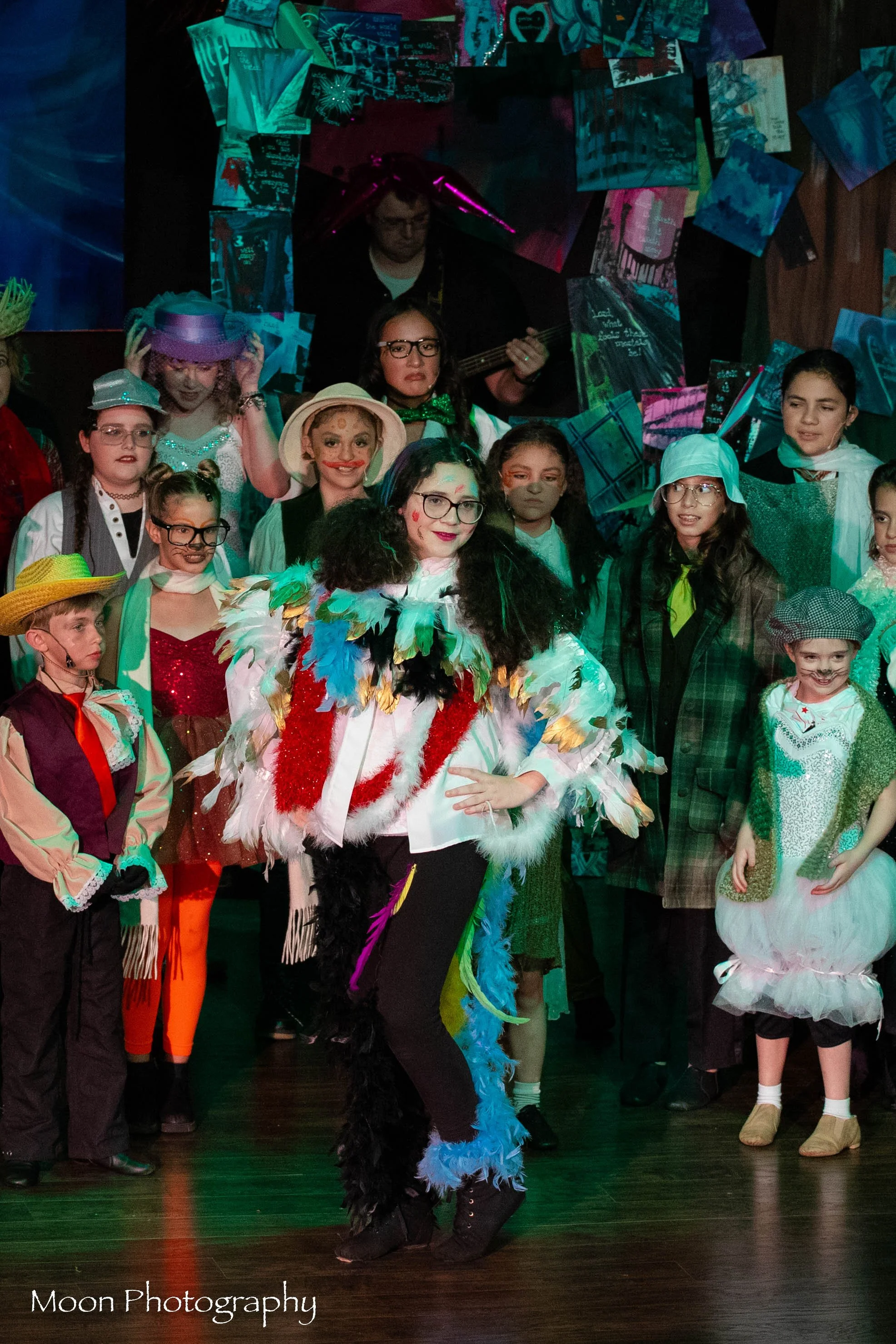Children and a woman in colorful costumes on stage during a theatrical performance, with a background decorated with books and props. A musician is playing guitar in the background.
