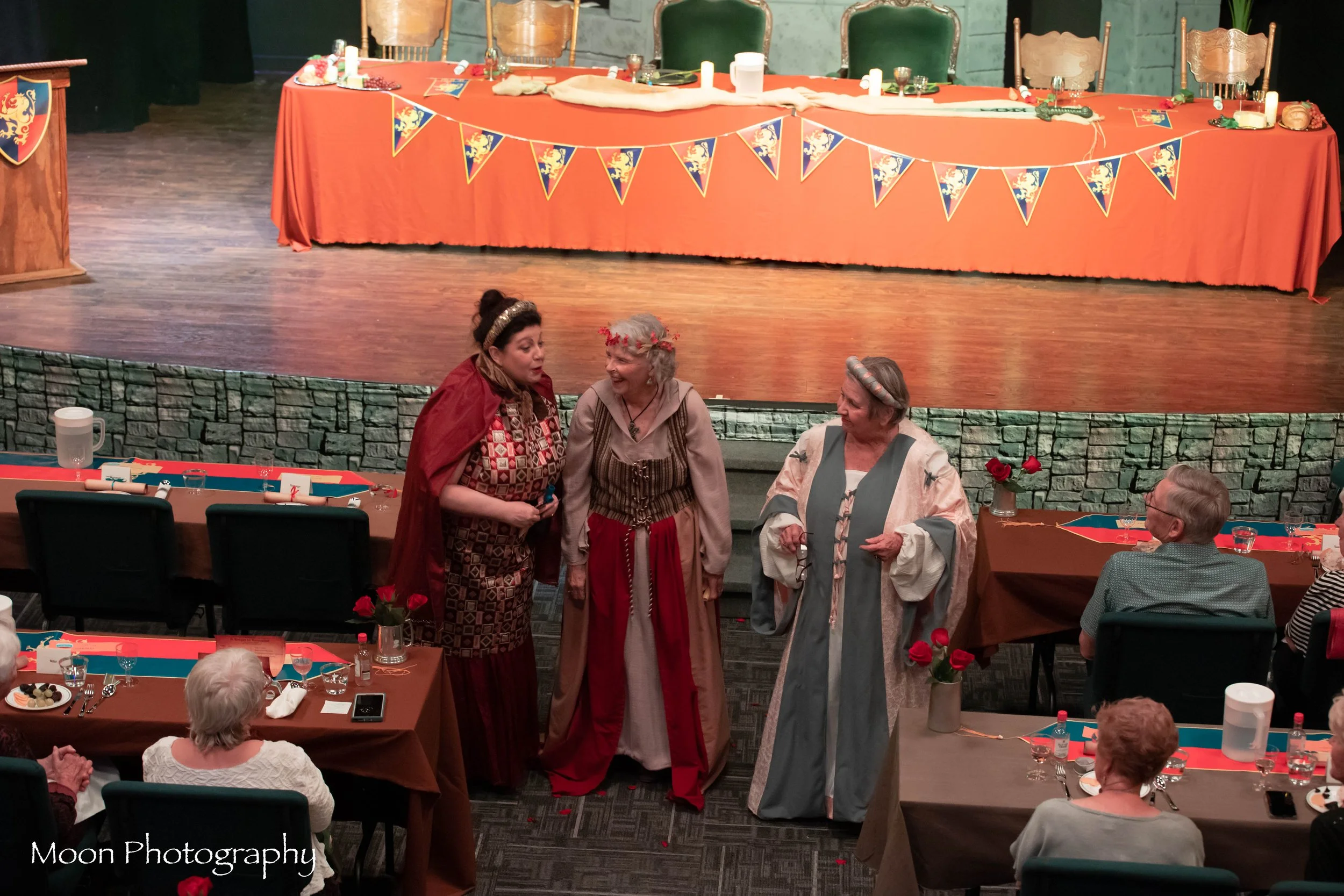 Three women dressed in medieval or fantasy costumes stand and speak on a stage at a banquet or themed event, with several seated attendees at tables decorated with red and blue tablecloths, flowers, and candles in a banquet hall.