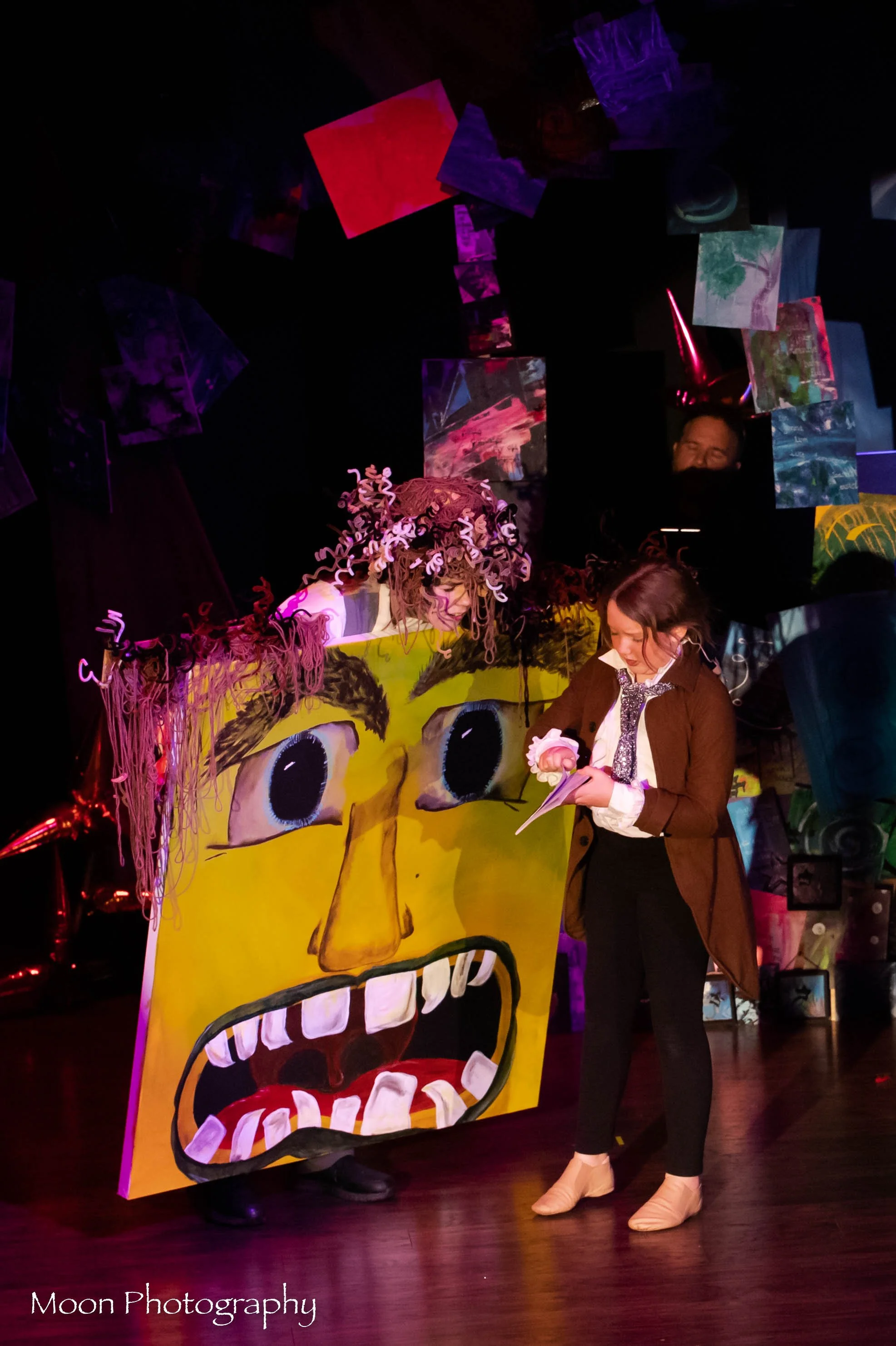 A woman stands next to a large colorful puppet with a yellow face, big blue eyes, and an open mouth showing teeth, with curly pink yarn hair. Another woman dressed as a character with a pink curly wig and a unicorn horn is behind the puppet. The back