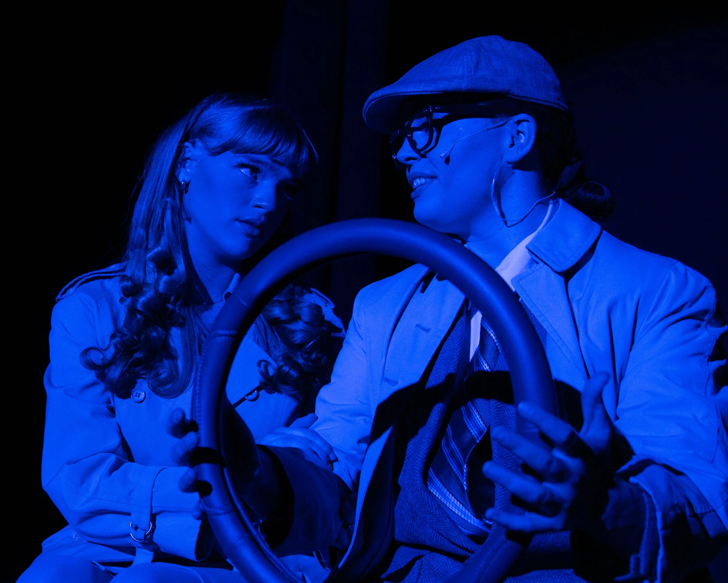 Two women engaged in a theatrical performance, illuminated with blue lighting, one holding a steering wheel prop.