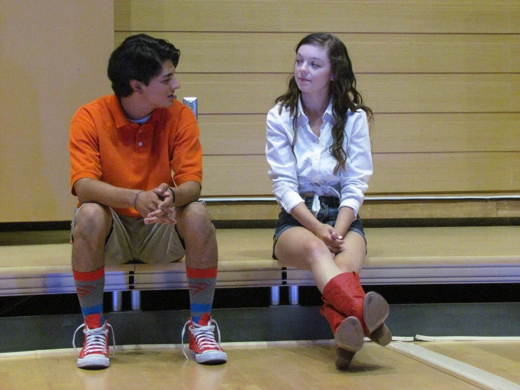 A teenage boy and girl sitting on a bench, engaged in conversation in a room with wooden paneling, the girl is wearing a white shirt and boots, and the boy is wearing an orange polo and khaki shorts.