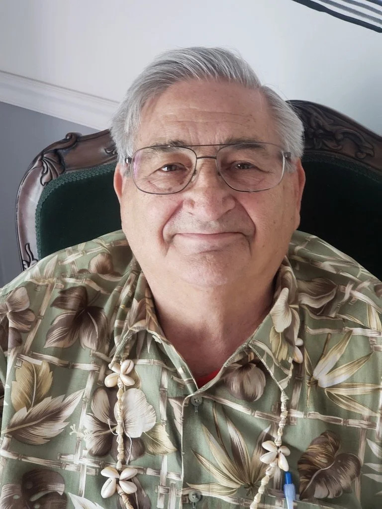 A smiling elderly man with gray hair and glasses, wearing a tropical floral shirt and a shell necklace, sitting in a green and dark wood armchair.
