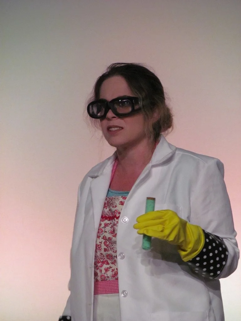 Woman dressed as a scientist wearing a white lab coat, large safety goggles, yellow rubber gloves, and holding a test tube. She has a confused or skeptical facial expression.