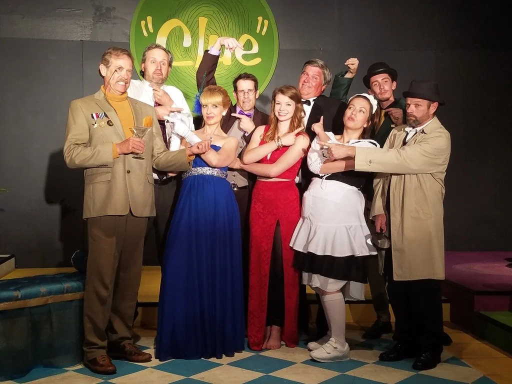 Group of nine people dressed in vintage costumes, posing on stage with a green circular sign that says 'Clue' in the background.
