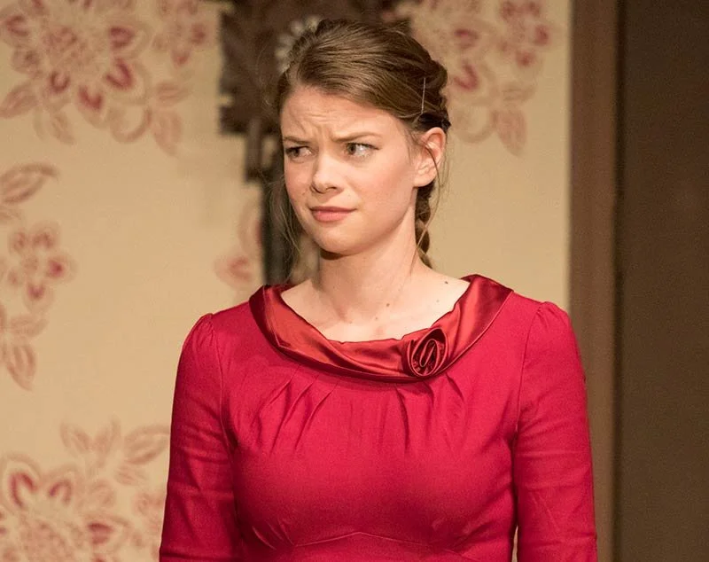 A woman with brown hair in a braid, wearing a red dress with a floral detail at the collar, has a confused or skeptical expression.