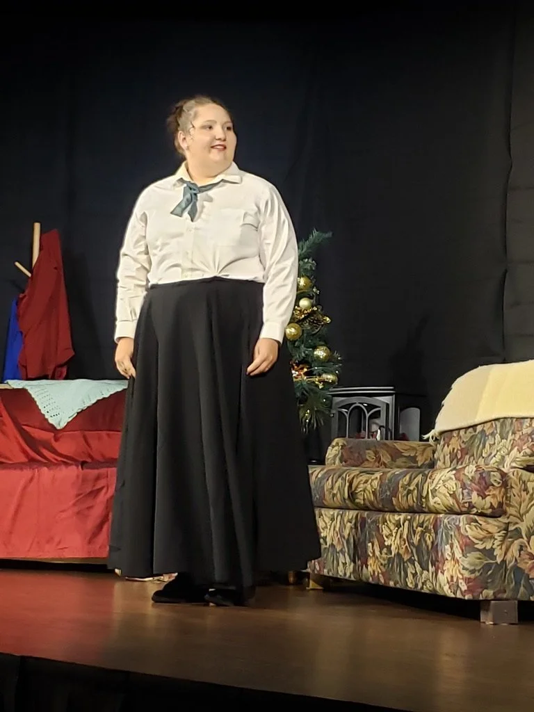A woman dressed in a white blouse and long black skirt standing on a stage with a Christmas tree and couches in the background.