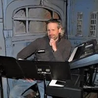 A man sitting at a keyboard and music stand, in front of a painted blue set with large windows, possibly during a rehearsal or performance.