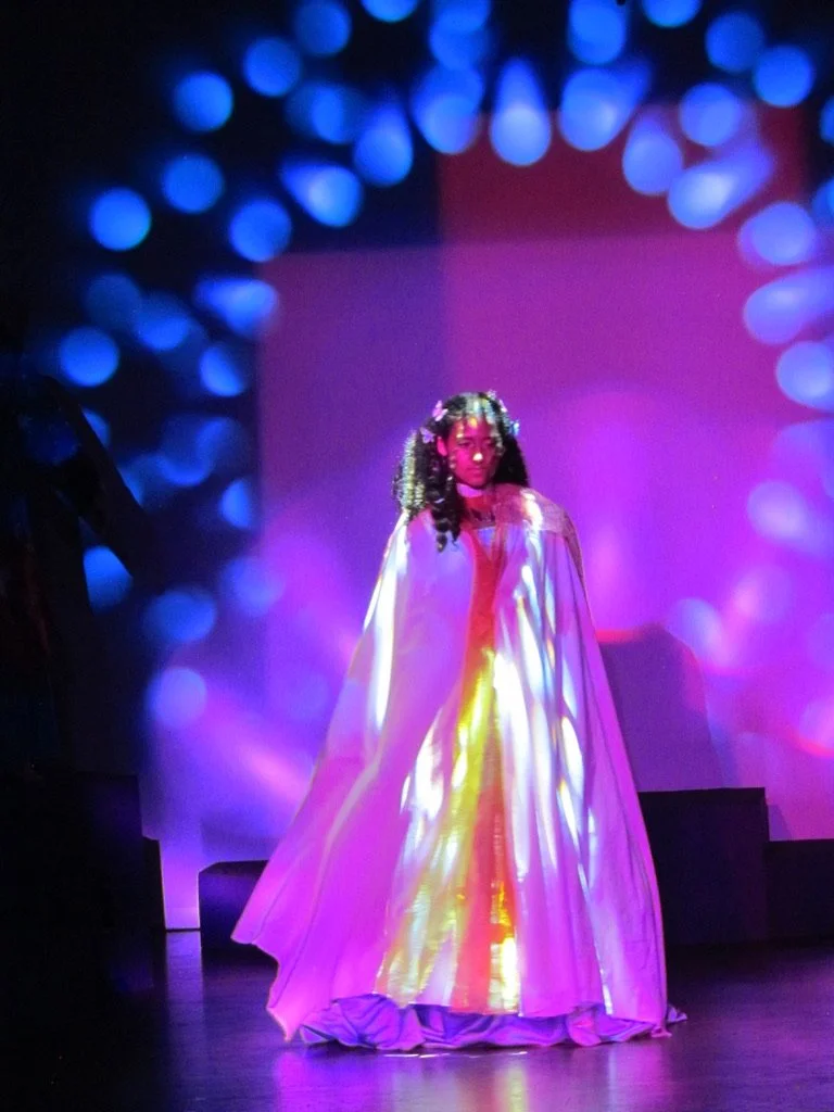 A person dressed in a flowing, multicolored gown with a glowing, colorful light projected onto it, standing on a stage with vibrant blue and purple bokeh lights in the background.