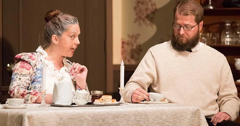 An older woman with gray hair in a bun, wearing a floral top, is gesturing animatedly while talking to a man with glasses and a beard, dressed in a beige sweater, sitting at a dining table with cups, a pitcher, and food.