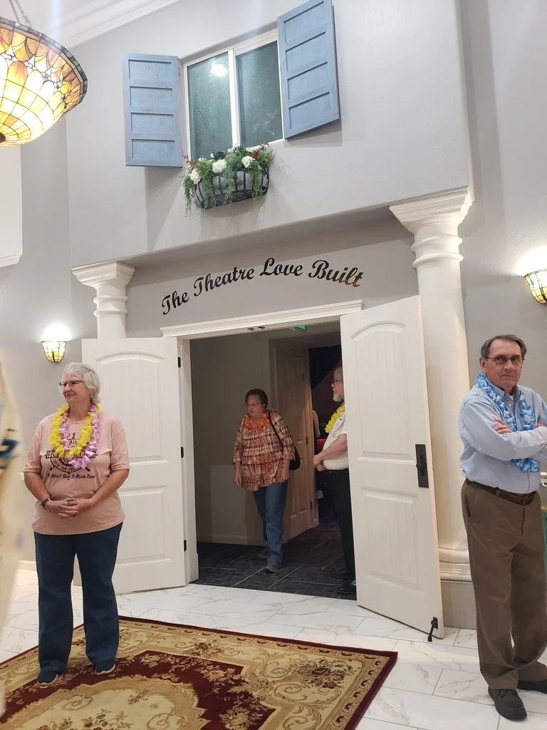 People standing in front of a theater entrance with a sign that reads 'The Theatre Love Built.' The room has white columns, open blue window shutters, and a floral arrangement outside the window. The floor is tiled, with a decorative rug near the ent