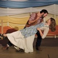 A man and woman dance together, with the man leaning over the woman who is leaning back, holding each other in a dance pose, in a decorated room with yellow, blue, and beige drapes.