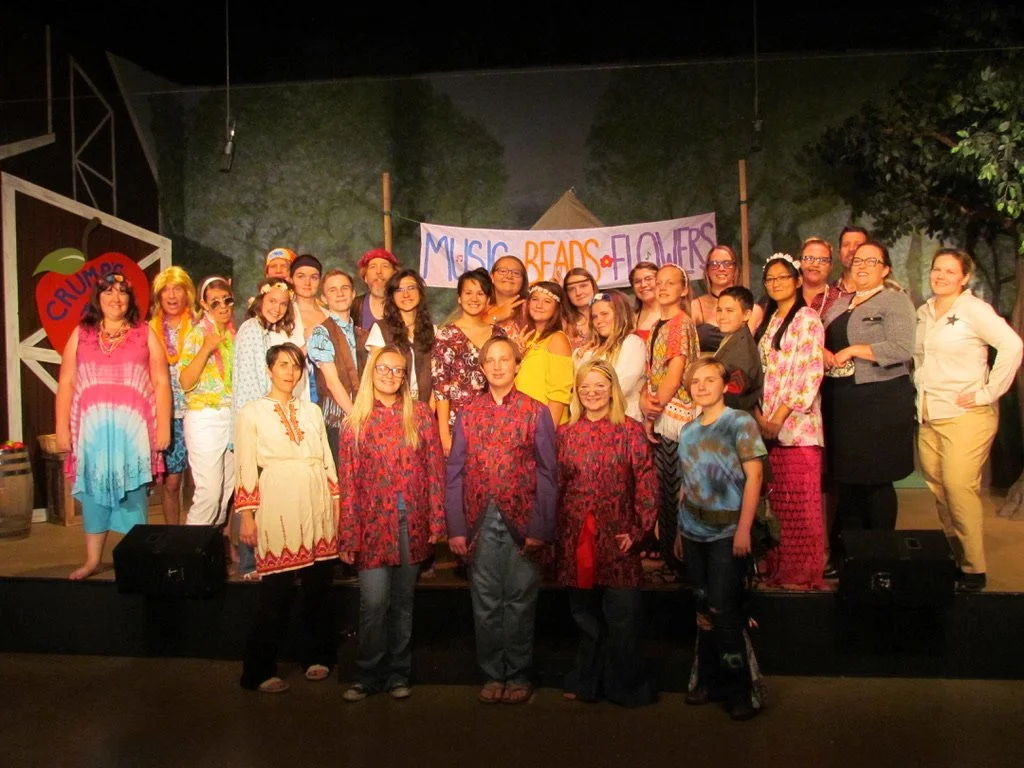 Group of children and adults on stage at a school play or performance in colorful costumes, with a sign in the background that says "Music, Beads, Flowers."