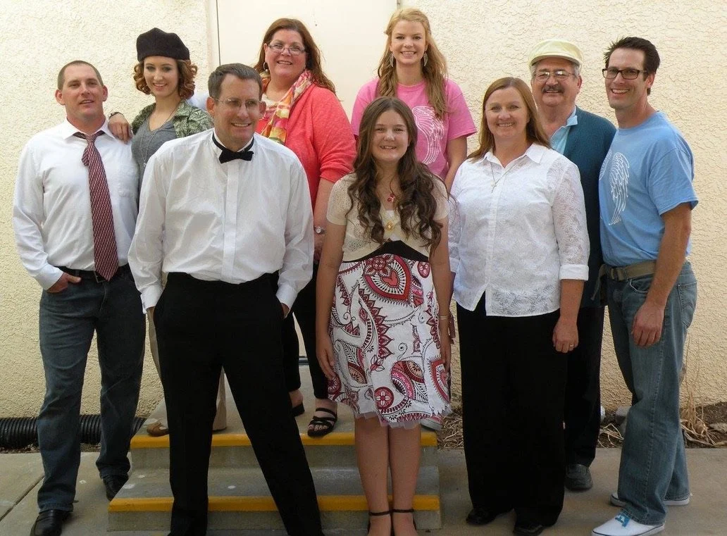Group of nine people standing on steps outside, smiling for the camera. They vary in age, wearing casual and semi-formal clothing, with some in glasses and a mix of genders and hair colors.