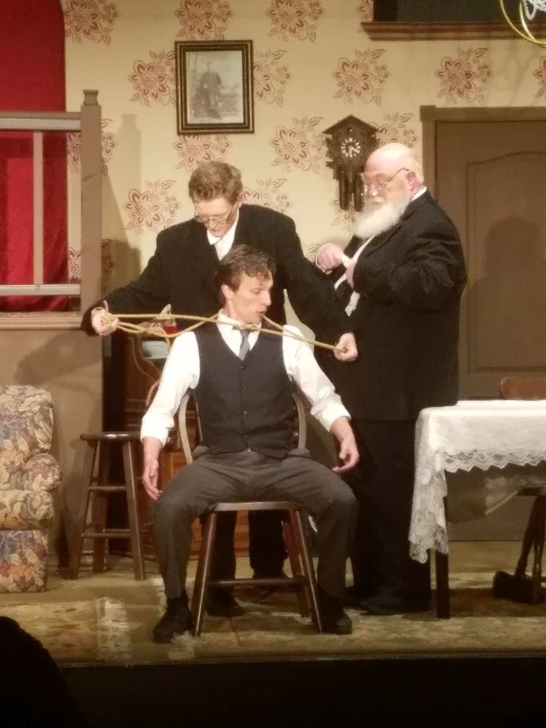 Three men on a stage involved in a theatrical scene. One man is sitting on a chair with a rope around his neck, while the other two stand behind him, one pulling the rope and the other holding a small whip. The setting looks like a vintage or old-fas