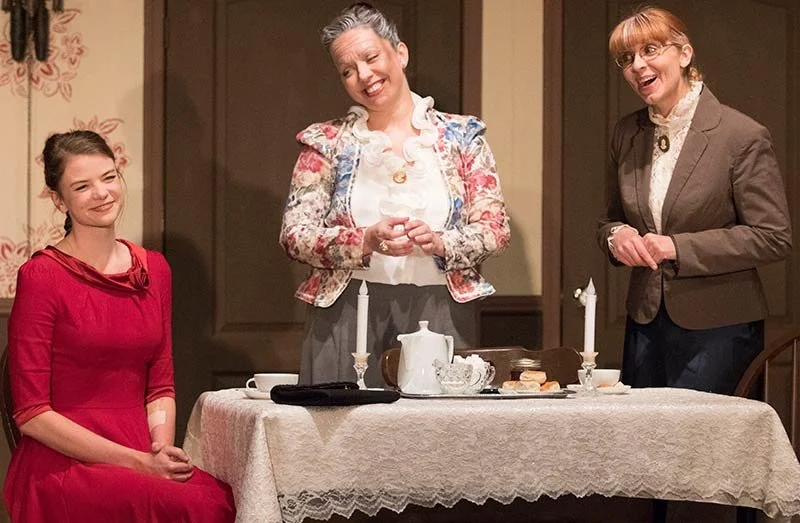 Three women around a dining table with a teapot, candles, and snacks, in a warmly lit room, possibly during a formal gathering or dinner.