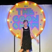 A person standing on stage in front of a sign that reads 'The Comic Show' with colorful lights around it.