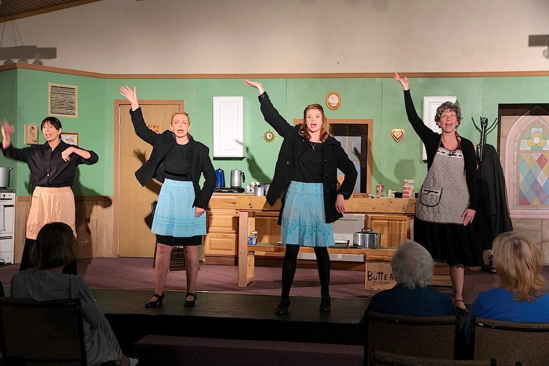 Four women performing on stage in a church basement kitchen set with audience members watching.