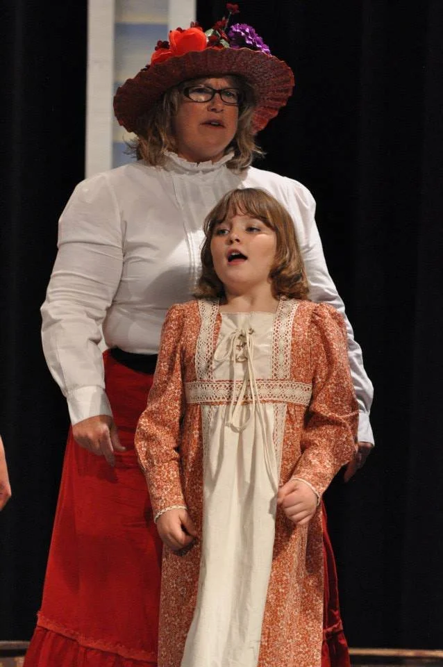 A woman and a girl performing on stage, with the woman wearing a large red hat decorated with flowers, white blouse, and red skirt, and the girl wearing a long patterned dress, both singing.
