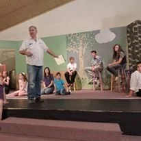A man performing on stage with children seated around him, some holding papers, in a decorated room with trees and clouds painted on the walls.