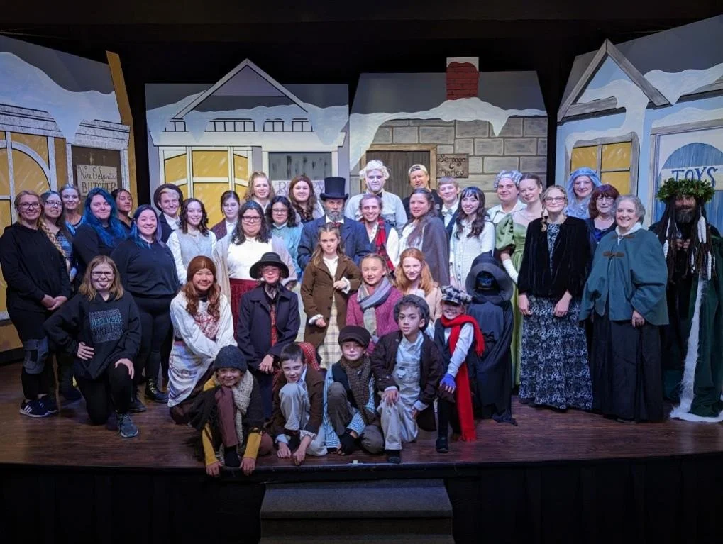 Group of children and adults on a stage, dressed in costumes for a theatrical production, with cartoon-like house backgrounds and snow accents.