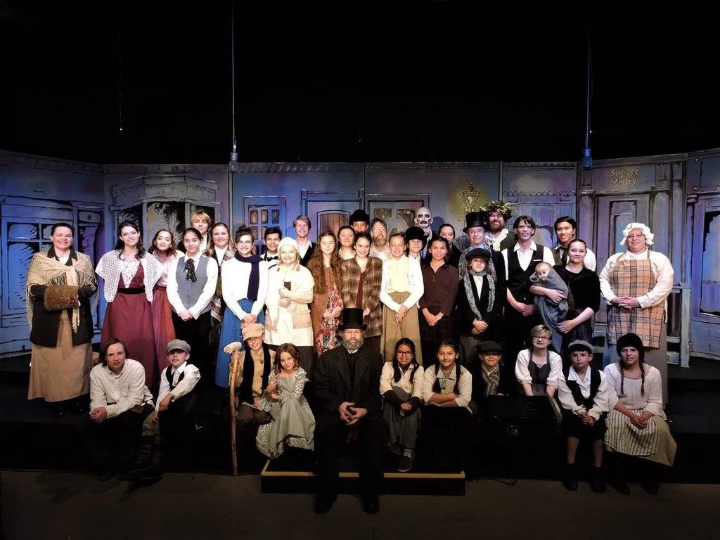A group of children and adults in period costumes posing on a theater stage for a cast photo.