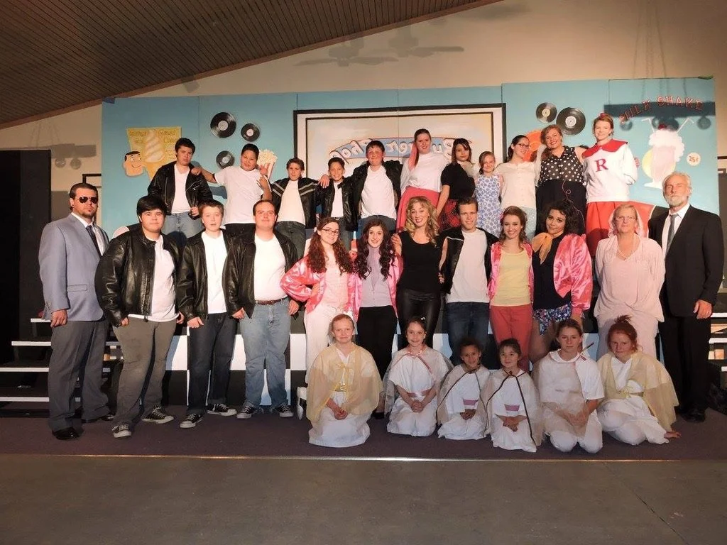 Group of children and adults posing on stage after a performance, with a colorful cartoon-themed backdrop and musical note decorations.