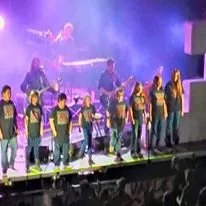 A group of children on stage performing a dance routine, with a singer in the background and stage fog creating a theatrical atmosphere.
