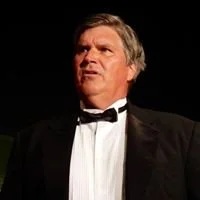 A man wearing a black tuxedo and bow tie, with a serious expression.